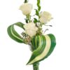Three white roses rise above slender greenery stems in a tall, clear glass vase. A large, variegated leaf loops gracefully around the blooms, framing small white filler flowers nestled at the base.