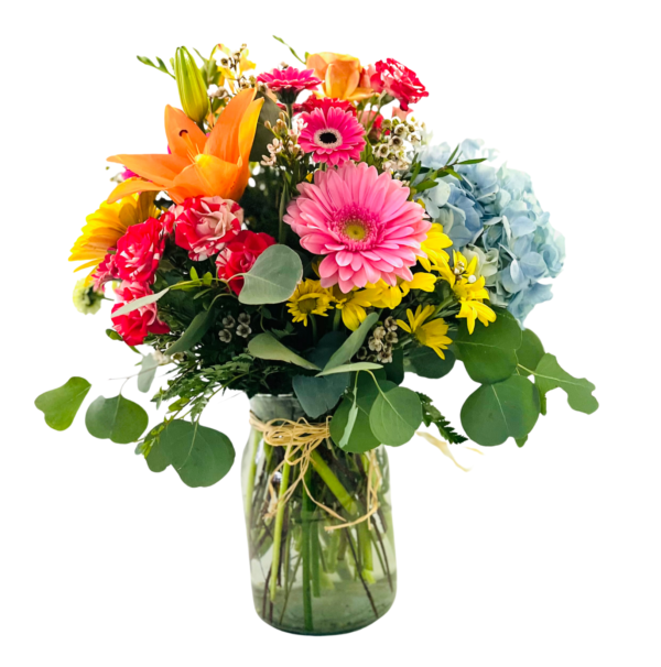 A tall glass vase tied with raffia holds a mixed bouquet featuring a pink gerbera center, an orange lily, red carnations, yellow daisies, pink waxflowers, and a cluster of blue hydrangea. Eucalyptus and fern fronds fan out around the blooms, with long eucalyptus stems draping over the vase’s rim. The arrangement bursts with bright, contrasting colors and lush greenery.