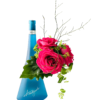 A frosted blue, triangular bottle labeled “Avantgarde” serves as a vase on the left. To the right, a cluster of vibrant hot pink roses nestles against a large glossy green leaf, accented by delicate green foliage and wispy, bare twig stems extending upward.