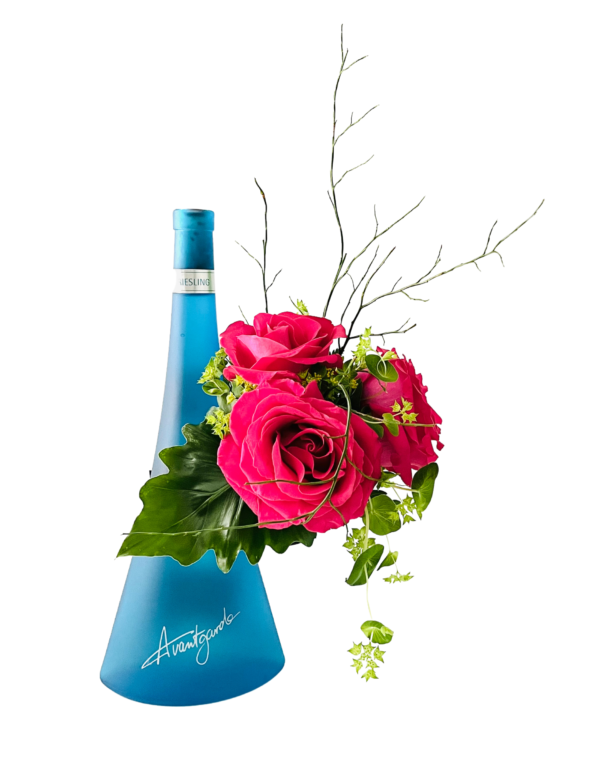 A frosted blue, triangular bottle labeled “Avantgarde” serves as a vase on the left. To the right, a cluster of vibrant hot pink roses nestles against a large glossy green leaf, accented by delicate green foliage and wispy, bare twig stems extending upward.