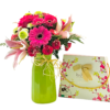 A tall, glossy lime-green vase tied with a raffia bow holds a lush mixed bouquet of pink lilies, hot pink gerbera daisies, red spray roses, chartreuse button mums, and small pink carnations nestled among fern and eucalyptus foliage. To the right of the vase, a rectangular La Stella chocolate gift box decorated with pink floral artwork and a gold ribbon handle leans against the vase.