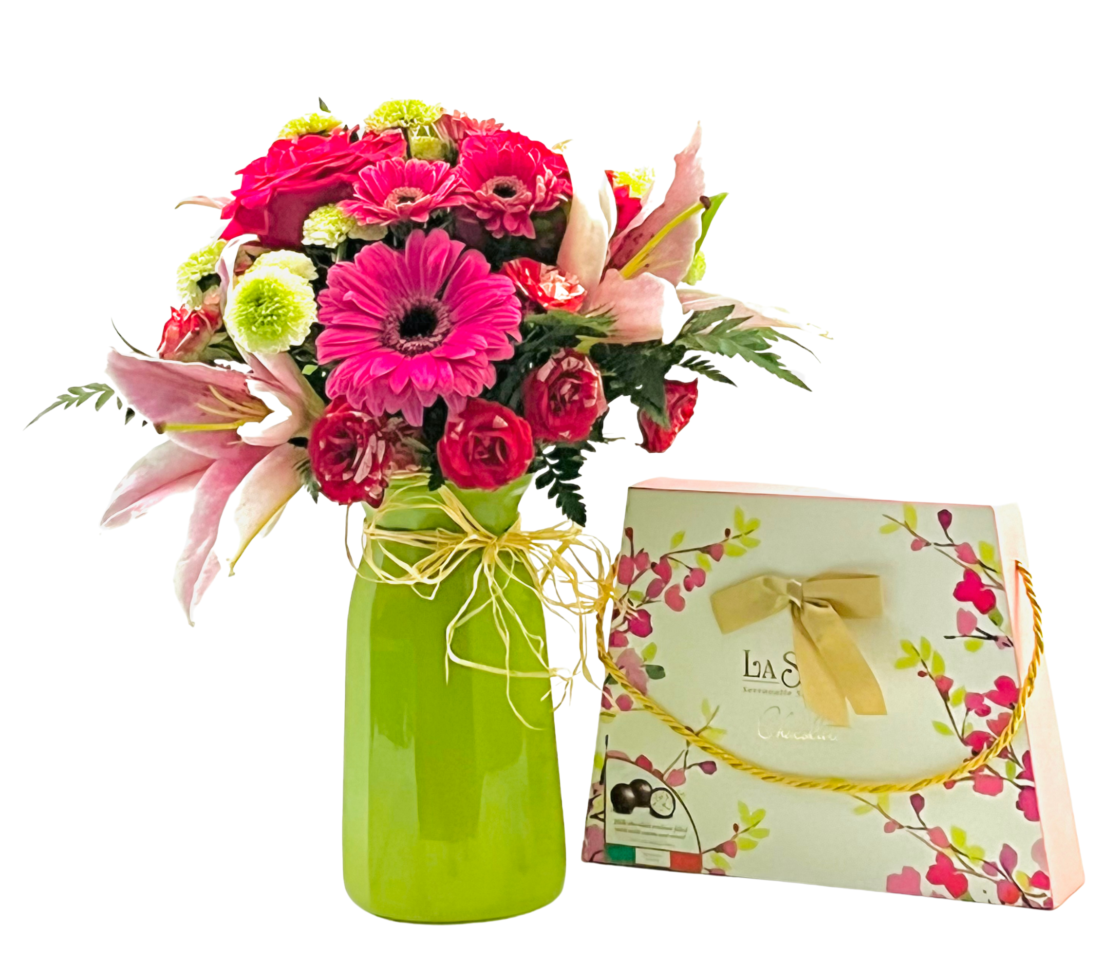 A tall, glossy lime-green vase tied with a raffia bow holds a lush mixed bouquet of pink lilies, hot pink gerbera daisies, red spray roses, chartreuse button mums, and small pink carnations nestled among fern and eucalyptus foliage. To the right of the vase, a rectangular La Stella chocolate gift box decorated with pink floral artwork and a gold ribbon handle leans against the vase.