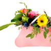 A pink, purse-shaped container holds a vibrant mixed bouquet. A bright yellow gerbera daisy and a soft pink gerbera daisy peek out at center, accompanied by peach freesia blossoms and spiky blue thistle. Glossy, looped palm leaves and long, variegated blades extend dramatically to the left, while cascading eucalyptus and delicate yellow-green filler foliage spill over the right edge. The arrangement is tied together with a pale pink ribbon looped across the top, creating a cheerful, asymmetrical display.