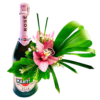 A small bottle of Martini Rosé holds a pink cymbidium orchid at its center. Glossy green leaves cradle the orchid, while tiny white waxflower buds cluster around it. A looping palm blade arcs to the right, and a few bright green grass blades rise behind, creating a sleek, modern arrangement on the bottle.