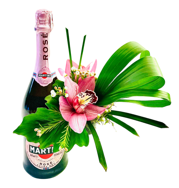 A small bottle of Martini Rosé holds a pink cymbidium orchid at its center. Glossy green leaves cradle the orchid, while tiny white waxflower buds cluster around it. A looping palm blade arcs to the right, and a few bright green grass blades rise behind, creating a sleek, modern arrangement on the bottle.