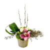 A small, rustic stone-textured pot holds a low, horizontally oriented arrangement. Central are three tall pussy willow branches with fuzzy catkins rising above a cluster of creamy white mini roses. To the right, feathery dried seeded branches extend outward, creating a wispy backdrop. At front left, a single pink orchid with a spotted burgundy-and-cream throat rests atop a bright green moss ball. Curved loops of glossy green ti leaf frame the left side, and tiny white waxflower buds and asparagus fern fill the spaces between, forming an elegant, asymmetrical design.