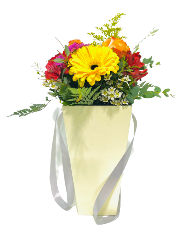 A pale yellow gift bag with silver ribbon handles holds a mixed floral arrangement: a bright yellow gerbera daisy at center, surrounded by orange rose buds, red alstroemeria, small pink blooms, white waxflowers, and assorted fern and eucalyptus foliage.