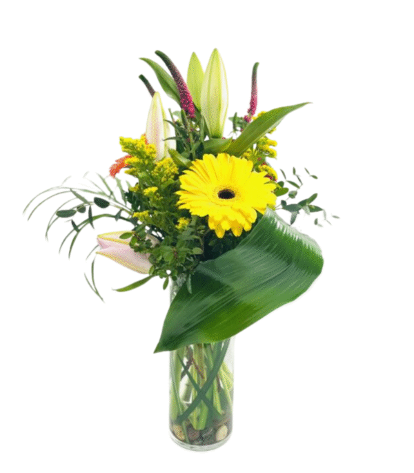 A tall, clear cylindrical vase filled with smooth river rocks holds a vibrant arrangement of flowers and greenery. At the center is a bright yellow gerbera daisy, surrounded by unopened pale pink lily buds, sprigs of goldenrod, long curved green leaves, and assorted leafy foliage, creating a modern, asymmetrical bouquet.