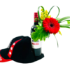 A tall bottle of 2020 Malbec wine stands upright, its neck adorned with a single bright red gerbera daisy, looping green palm leaf loops, a spray of small yellow blooms, and a large split green leaf tucked behind. In front of the bottle, a black baseball cap with a red brim and white “Lethbridge” lettering rests tipped over, featuring two red maple leaf graphics on its side panels.