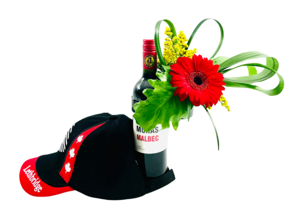 A tall bottle of 2020 Malbec wine stands upright, its neck adorned with a single bright red gerbera daisy, looping green palm leaf loops, a spray of small yellow blooms, and a large split green leaf tucked behind. In front of the bottle, a black baseball cap with a red brim and white “Lethbridge” lettering rests tipped over, featuring two red maple leaf graphics on its side panels.