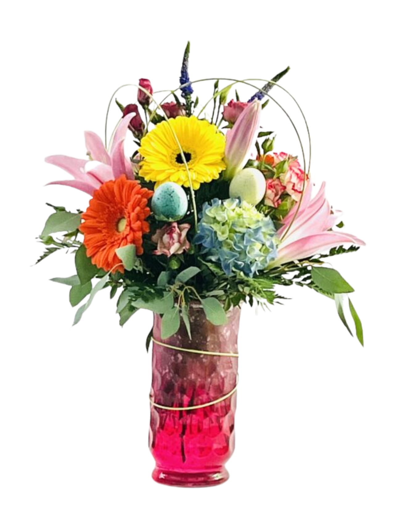 A tall, faceted magenta glass vase wrapped with a thin gold wire holds a springtime bouquet of mixed blooms and greenery. At the front, a bright orange gerbera daisy and a yellow gerbera daisy sit above a cluster of pale blue and green hydrangea petals. Pink Asiatic lilies open behind them, and a few unopened pink lily buds extend upward. Small speckled pastel eggs (light blue and pale yellow) are tucked among the foliage. A spiky blue thistle and deep red spray roses peek out toward the back, while fuzzy gray pussy willow catkins rise tall above all. Fern fronds, eucalyptus leaves, and slender green blades of grass fill in around the edges, creating a lush, textured arrangement.
