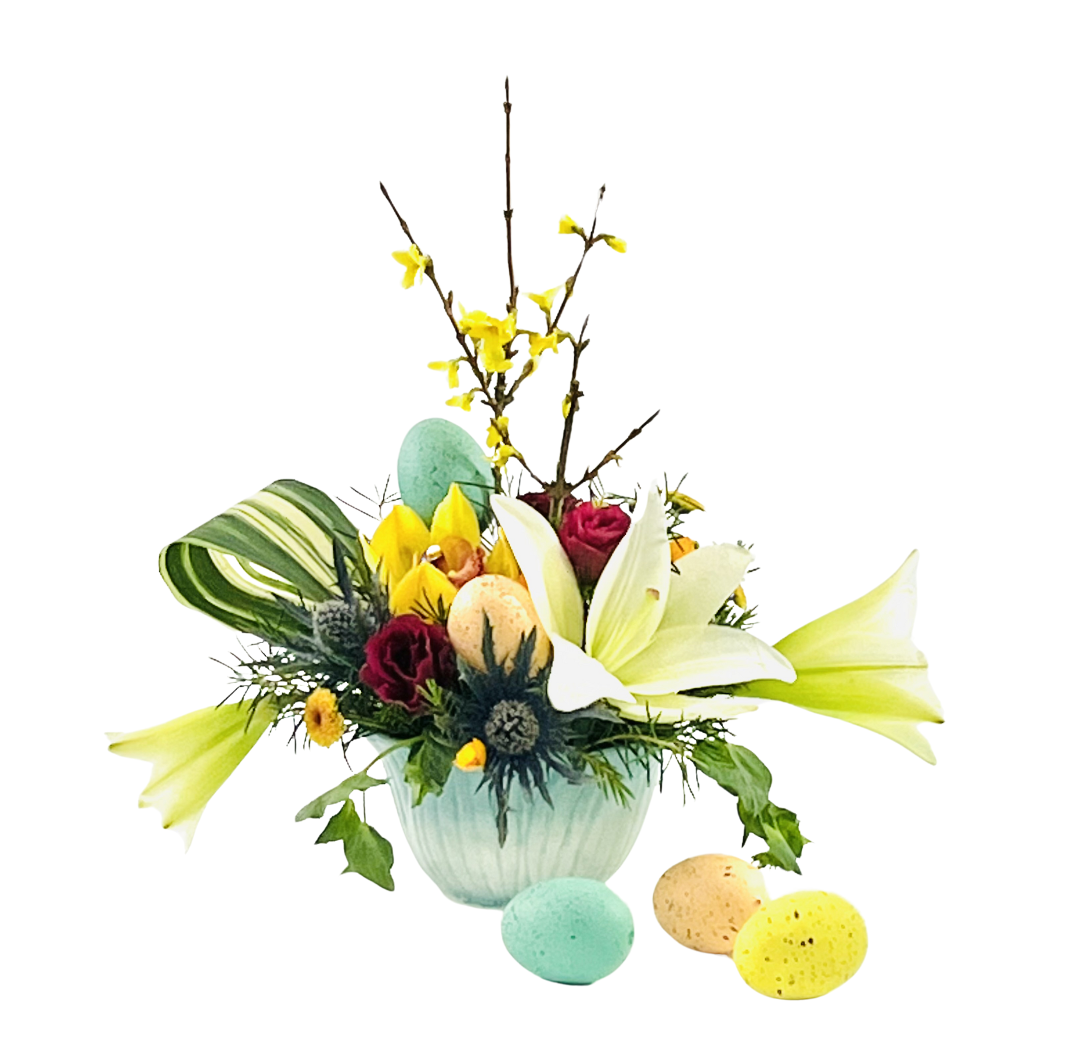 A shallow, ribbed aqua bowl holds a spring arrangement of mixed blooms and branches. Two pale green amaryllis blooms extend wide on the right and left, framing bright yellow orchids and deep red spray roses clustered at center. Spiky blue thistle, small yellow button mums, and feathery greenery fill in around the base. Tall forsythia branches with yellow blossoms rise behind, and a variegated palm leaf curls to the left. Three pastel-speckled Easter eggs (mint, peach, and yellow) nestle among the flowers and trail in front on the table.
