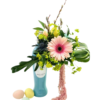 A turquoise Pinot Grigio bottle repurposed as a vase holds an arrangement of spring blooms and greenery. A large pale pink gerbera daisy sits front and center, surrounded by spiky blue thistle, bright chartreuse Lady’s Mantle, and long curved palm fronds. Three pussy willow stems extend upward behind a pair of speckled pastel eggs tucked among the foliage. A trailing strand of soft pink amaranthus drapes down the bottle’s side, and three additional pastel-speckled eggs (mint green, pale yellow, and peach) rest at the bottle’s base.