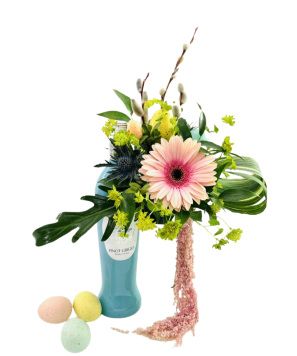 A turquoise Pinot Grigio bottle repurposed as a vase holds an arrangement of spring blooms and greenery. A large pale pink gerbera daisy sits front and center, surrounded by spiky blue thistle, bright chartreuse Lady’s Mantle, and long curved palm fronds. Three pussy willow stems extend upward behind a pair of speckled pastel eggs tucked among the foliage. A trailing strand of soft pink amaranthus drapes down the bottle’s side, and three additional pastel-speckled eggs (mint green, pale yellow, and peach) rest at the bottle’s base.