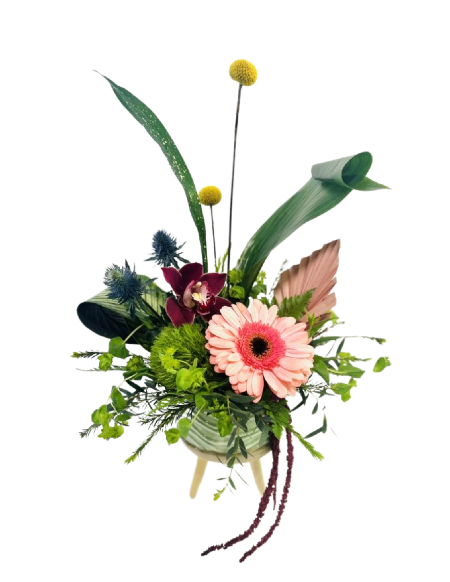 A low, round green ceramic vase on three small wooden legs contains an eclectic arrangement: a soft pink gerbera daisy at front right, a deep burgundy orchid nestled behind, bright yellow billy ball stems rising above, spiky blue thistle blooms to the left, a lime-green globe dianthus at center, and assorted greenery—including long curved blades of palm leaf, a dried blush-pink palm fan, and trailing burgundy amaranthus—creating a dynamic, textured bouquet.