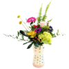 A ceramic vase shaped like a stylized smiling face—with closed eyes, rosy cheeks, and a polka-dotted base—holds a vibrant, mixed bouquet. Fern fronds rise tall behind a cluster of blooms including a bright yellow orchid, a pink gerbera daisy, a pale pink rose, a purple anemone, and a chartreuse hydrangea. Two round yellow billy balls on slender stems extend upward at left. Variegated palm leaves curve dramatically to one side, while eucalyptus and glossy foliage spill gracefully over the vase’s rim, creating a whimsical, colorful arrangement.