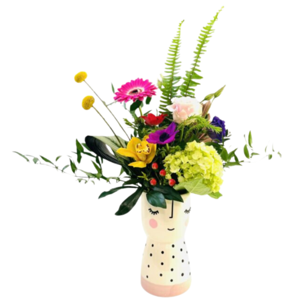A ceramic vase shaped like a stylized smiling face—with closed eyes, rosy cheeks, and a polka-dotted base—holds a vibrant, mixed bouquet. Fern fronds rise tall behind a cluster of blooms including a bright yellow orchid, a pink gerbera daisy, a pale pink rose, a purple anemone, and a chartreuse hydrangea. Two round yellow billy balls on slender stems extend upward at left. Variegated palm leaves curve dramatically to one side, while eucalyptus and glossy foliage spill gracefully over the vase’s rim, creating a whimsical, colorful arrangement.