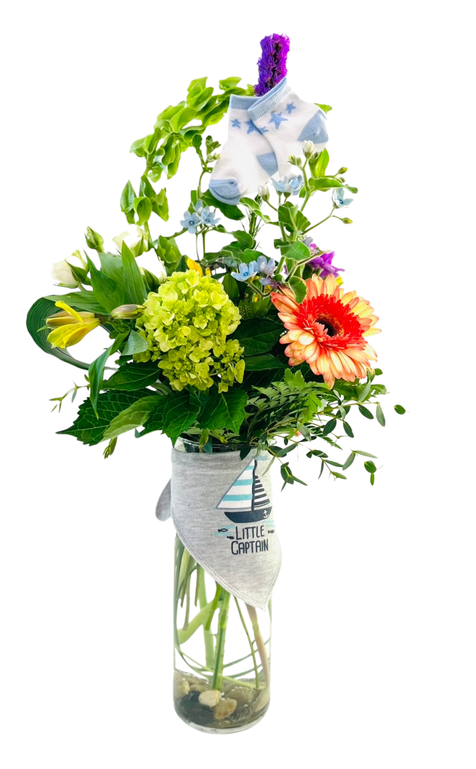 A tall, clear glass cylinder vase partially filled with pebbles holds a celebratory mixed bouquet. Fern fronds, eucalyptus, and variegated palm leaves form a lush green backdrop. A cluster of bright chartreuse hydrangea sits at center, alongside an orange-pink gerbera daisy and yellow alstroemeria blooms. Sprays of tiny blue forget-me-nots and purple liatris spikes rise above, with trailing ivy vines weaving through the greenery. Draped around the vase’s neck is a gray bib printed with a sailboat graphic and the words “Little Captain.” At the very top, a pair of white baby socks with light blue accents and star patterns are tucked among the stems, creating a whimsical, new-baby accent.