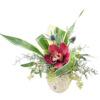 A mottled stone-textured round vase holds an asymmetrical arrangement centered on a single deep burgundy orchid with a spotted cream-and-burgundy throat. It is surrounded by spiky blue thistle blooms, sprays of silvery cedar, and small clusters of green buds layered among eucalyptus stems. Broad, variegated palm leaves curve dramatically to the left and right, framing the orchid and adding a sweeping, sculptural element to the composition.
