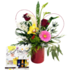 A glossy red cylindrical vase holds a lush mixed bouquet. Three deep red roses rise on tall stems amid feathery pampas grass and a speckled green ti leaf. In front, a bright yellow orchid and a vivid pink gerbera daisy sit beside spiky blue thistle and a mossy green dianthus ball. Wisps of fern and cascading eucalyptus fill out the arrangement, while two red wire loops form subtle heart shapes behind the flowers. To the left of the vase, a “La Dolce Vita” chocolate box in white packaging displays miniature liqueur-filled chocolates in metallic blue, orange, and gold wrappers.