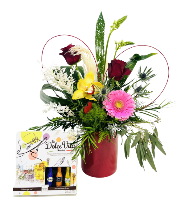 A glossy red cylindrical vase holds a lush mixed bouquet. Three deep red roses rise on tall stems amid feathery pampas grass and a speckled green ti leaf. In front, a bright yellow orchid and a vivid pink gerbera daisy sit beside spiky blue thistle and a mossy green dianthus ball. Wisps of fern and cascading eucalyptus fill out the arrangement, while two red wire loops form subtle heart shapes behind the flowers. To the left of the vase, a “La Dolce Vita” chocolate box in white packaging displays miniature liqueur-filled chocolates in metallic blue, orange, and gold wrappers.