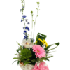 A white, perforated ceramic elephant-shaped pot holds a spring bouquet of mixed blooms and greenery. At the base, a bright green dianthus moss ball and delicate fern fronds fill out the arrangement. In front, a pale pink gerbera daisy and a white-and-pink cymbidium orchid sit among glossy variegated palm leaves and small yellow button chrysanthemums. Tall royal blue larkspur stems rise behind, flanked by slender leafy branches and an upright green lily bud. Draped over the front of the elephant is a pair of tiny pink-and-white baby socks with a heart pattern, creating a playful, celebratory touch.