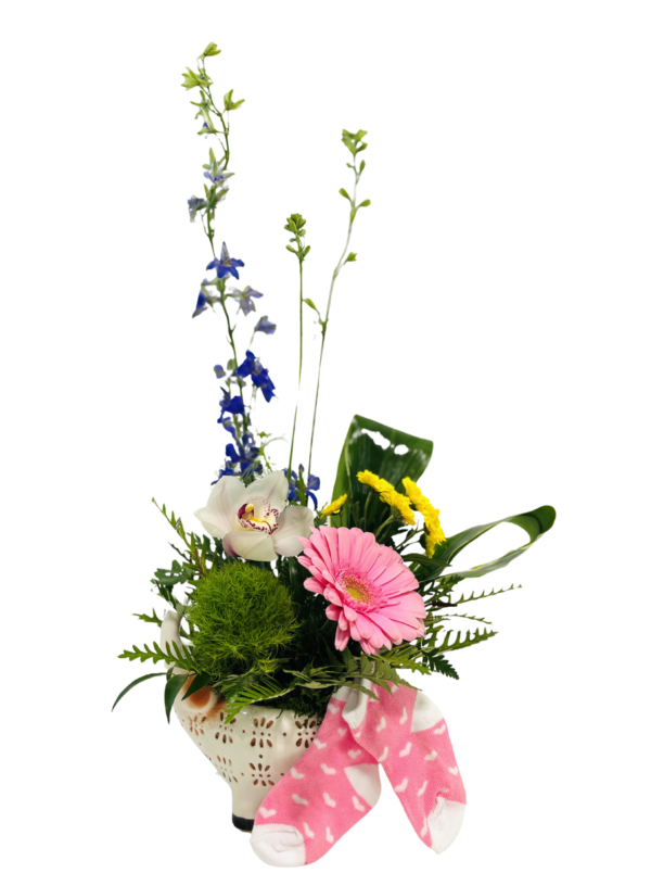 A white, perforated ceramic elephant-shaped pot holds a spring bouquet of mixed blooms and greenery. At the base, a bright green dianthus moss ball and delicate fern fronds fill out the arrangement. In front, a pale pink gerbera daisy and a white-and-pink cymbidium orchid sit among glossy variegated palm leaves and small yellow button chrysanthemums. Tall royal blue larkspur stems rise behind, flanked by slender leafy branches and an upright green lily bud. Draped over the front of the elephant is a pair of tiny pink-and-white baby socks with a heart pattern, creating a playful, celebratory touch.