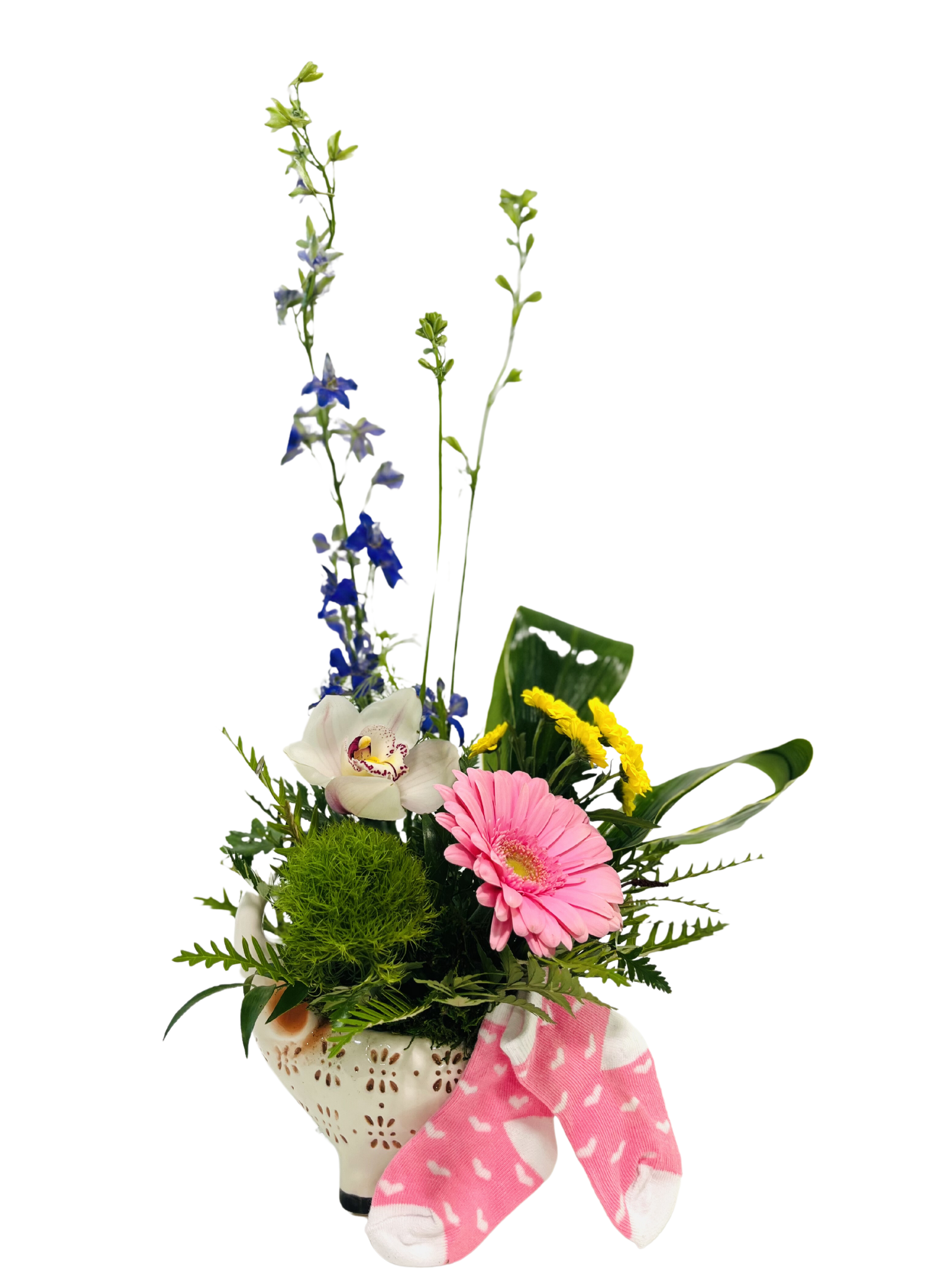 A white, perforated ceramic elephant-shaped pot holds a spring bouquet of mixed blooms and greenery. At the base, a bright green dianthus moss ball and delicate fern fronds fill out the arrangement. In front, a pale pink gerbera daisy and a white-and-pink cymbidium orchid sit among glossy variegated palm leaves and small yellow button chrysanthemums. Tall royal blue larkspur stems rise behind, flanked by slender leafy branches and an upright green lily bud. Draped over the front of the elephant is a pair of tiny pink-and-white baby socks with a heart pattern, creating a playful, celebratory touch.