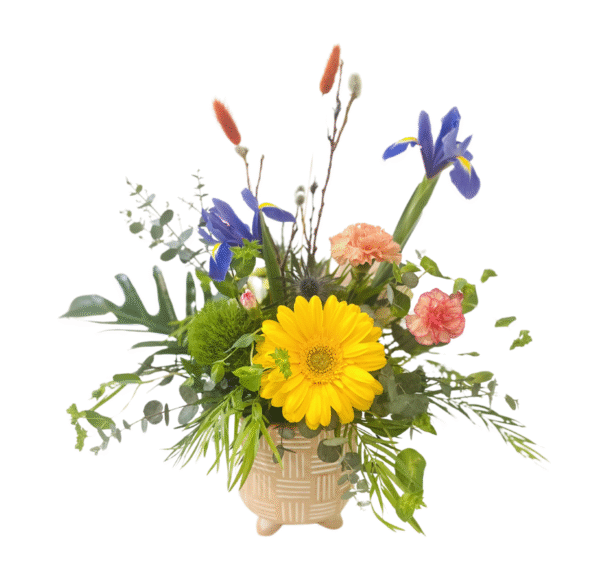 A beige ceramic pot on three short feet holds a lush, asymmetrical arrangement. A bright yellow gerbera daisy sits front and center, flanked by a fluffy green dianthus. Two deep blue iris bloom at upper right and left, their tall stems rising above sprigs of orange bunny tail grasses. A soft peach carnation and a pink-edged spray carnation tuck into the right side. Assorted greenery—including long palm fronds, eucalyptus, and delicate leafy foliage—flares outward, creating a full, natural silhouette.