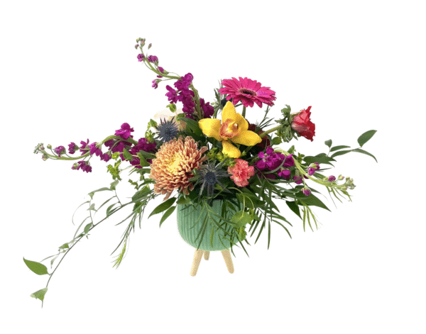 A green ribbed ceramic vase on three short wooden legs holds a sprawling arrangement of blooms and greenery. At center is a large peach chrysanthemum, flanked by a yellow orchid and a hot pink gerbera daisy. Magenta stock flowers cascade to the left, while blue thistle, coral carnations, small yellow daisies, and trailing eucalyptus leaves fill out the composition with a lush, asymmetrical silhouette.
