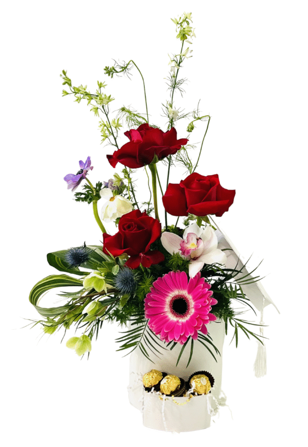 A tall white gift box with a hinged lid holds a theatrical floral arrangement and a pull‐out drawer of chocolates. At the base, the drawer is open to reveal three gold‐wrapped Ferrero Rocher chocolates nestled in white crinkle paper. Above, three long‐stemmed red roses rise vertically amid feathery asparagus fern and wispy white larkspur. Clustered below are a bright pink gerbera daisy, a white and pink orchid, and spiky blue thistle surrounded by glossy green leaves, curly palm fronds, and small green hellebore blooms, creating a layered, elegant presentation.