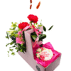 A pink cardboard gift box holds a mixed floral arrangement in the top compartment and a decorated cookie in the lower compartment. The flowers include bright red carnations, a pink gerbera daisy, yellow spray carnations, pink orchids, blue thistle accents, and greenery, tied with a pink “Mother’s Day” ribbon. In the lower section, a round iced sugar cookie rests on pink tissue paper, decorated with piped flowers and the words “Happy Mother’s Day.”