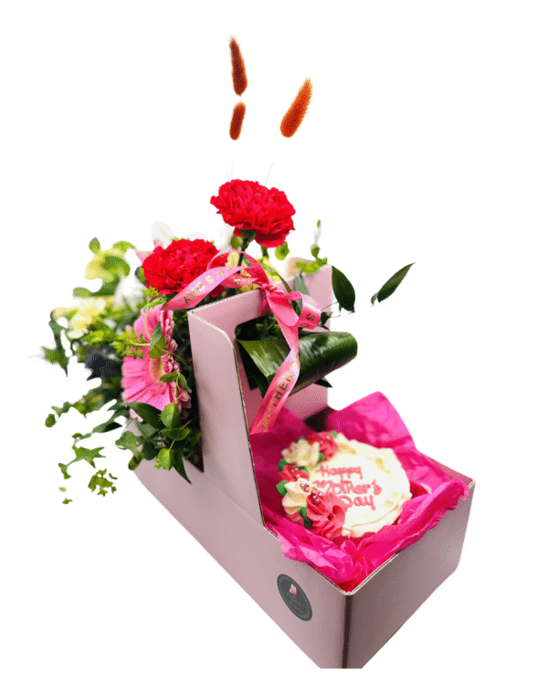 A pink cardboard gift box holds a mixed floral arrangement in the top compartment and a decorated cookie in the lower compartment. The flowers include bright red carnations, a pink gerbera daisy, yellow spray carnations, pink orchids, blue thistle accents, and greenery, tied with a pink “Mother’s Day” ribbon. In the lower section, a round iced sugar cookie rests on pink tissue paper, decorated with piped flowers and the words “Happy Mother’s Day.”