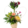 A textured beige ceramic pot on three short feet holds an eclectic arrangement: tall red alstroemeria stalks rise at center, surrounded by a peach carnation, small hot pink spray carnations, and a large golden-yellow chrysanthemum. Bright yellow daisy-like blooms and a fuzzy green dianthus add pops of color, while assorted greenery—including glossy leaves, fern fronds, and tiny white waxflowers—fills out the base. Two looping pale grass blades form a subtle heart shape behind the flowers, and a small wooden red heart pick accents the central greenery.