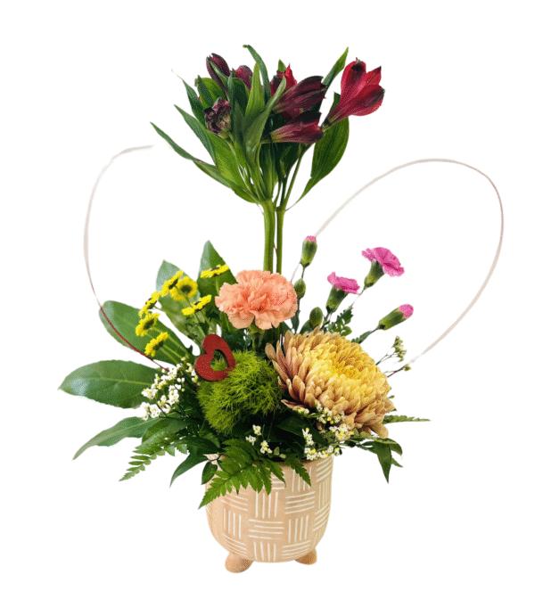 A textured beige ceramic pot on three short feet holds an eclectic arrangement: tall red alstroemeria stalks rise at center, surrounded by a peach carnation, small hot pink spray carnations, and a large golden-yellow chrysanthemum. Bright yellow daisy-like blooms and a fuzzy green dianthus add pops of color, while assorted greenery—including glossy leaves, fern fronds, and tiny white waxflowers—fills out the base. Two looping pale grass blades form a subtle heart shape behind the flowers, and a small wooden red heart pick accents the central greenery.