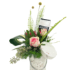 A decorative cylindrical gift box with a lid askew holds a lush floral arrangement of pink roses, red protea, assorted greenery (fern fronds, curled ti leaf), and small berries, accented by two yellow billy ball stems and a Lindt “Cranberry & Nuts” chocolate bar tucked among the blooms.