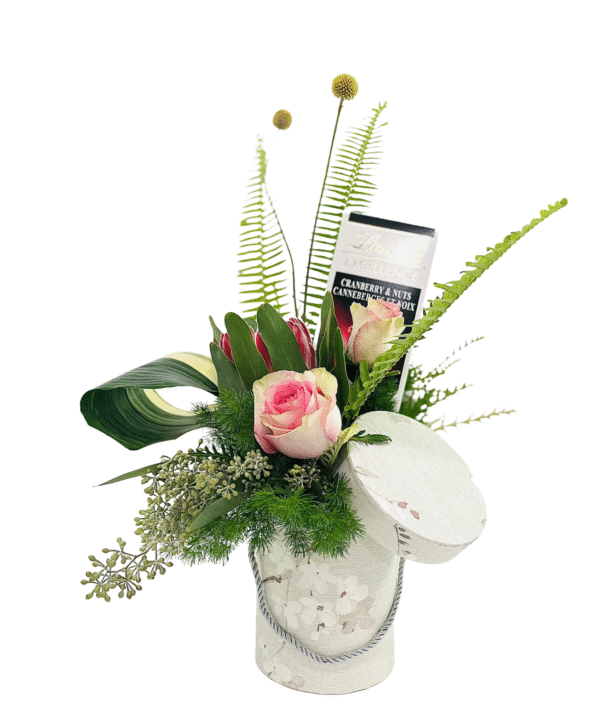 A decorative cylindrical gift box with a lid askew holds a lush floral arrangement of pink roses, red protea, assorted greenery (fern fronds, curled ti leaf), and small berries, accented by two yellow billy ball stems and a Lindt “Cranberry & Nuts” chocolate bar tucked among the blooms.