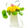A white gift bag with long ribbon handles holds a small arrangement of three vivid yellow roses topped by a single pink orchid bloom. Rounded eucalyptus leaves fan out around the roses, and two loops of decorative green grass rise from the back, creating a bright, modern floral display.