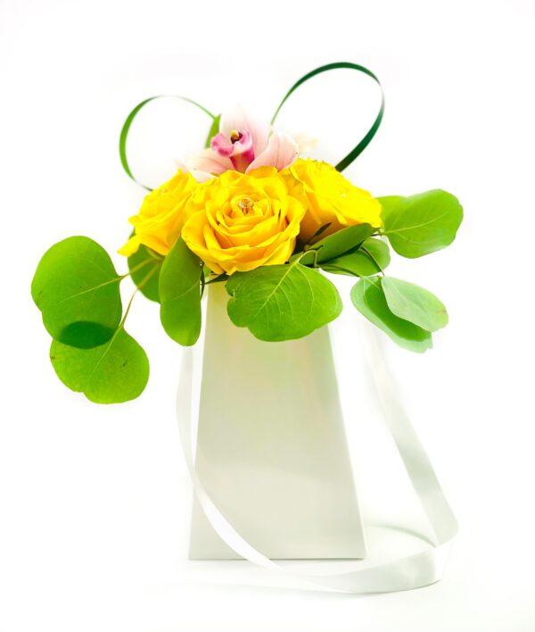 A white gift bag with long ribbon handles holds a small arrangement of three vivid yellow roses topped by a single pink orchid bloom. Rounded eucalyptus leaves fan out around the roses, and two loops of decorative green grass rise from the back, creating a bright, modern floral display.